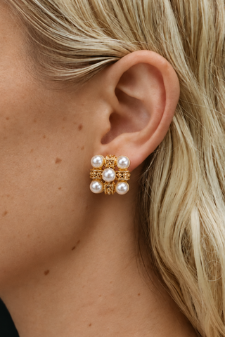 A close-up of a model's ear wearing a gold and pearl stud earring. The earring features a gold geometric frame with six white pearl-like beads.