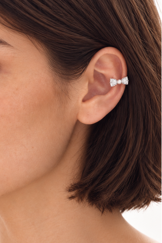 A close-up of a woman's ear wearing a small silver ear cuff with three clear gemstones, set against a clean white background.