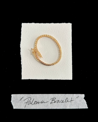 A gold-toned bangle bracelet with a textured, woven pattern and a small bow detail at the front, displayed on a white textured card with a handwritten label below reading 'Paloma Bracelet' against a solid black background.