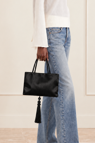 A model wearing a white long-sleeve mock-neck top, light-wash wide-leg jeans, and black pointed flats, holding a black leather handbag with a tassel detail against a cream paneled wall.