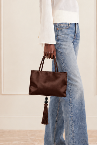 A model wearing a white long-sleeve mock-neck top, light-wash wide-leg jeans, and black pointed flats, holding a dark brown leather handbag with a tassel.